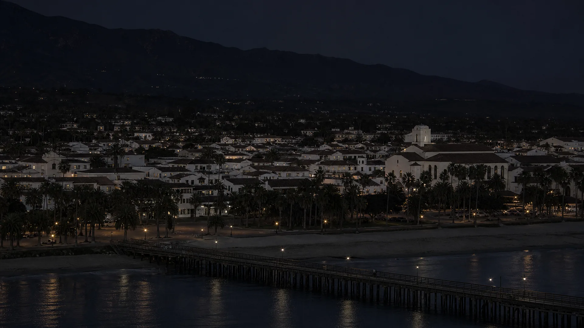 An elevated pre-dawn view over Santa Barbara's Spanish Colonial rooftops, waterfront, Stearns Wharf, and dark mountain backdrop.