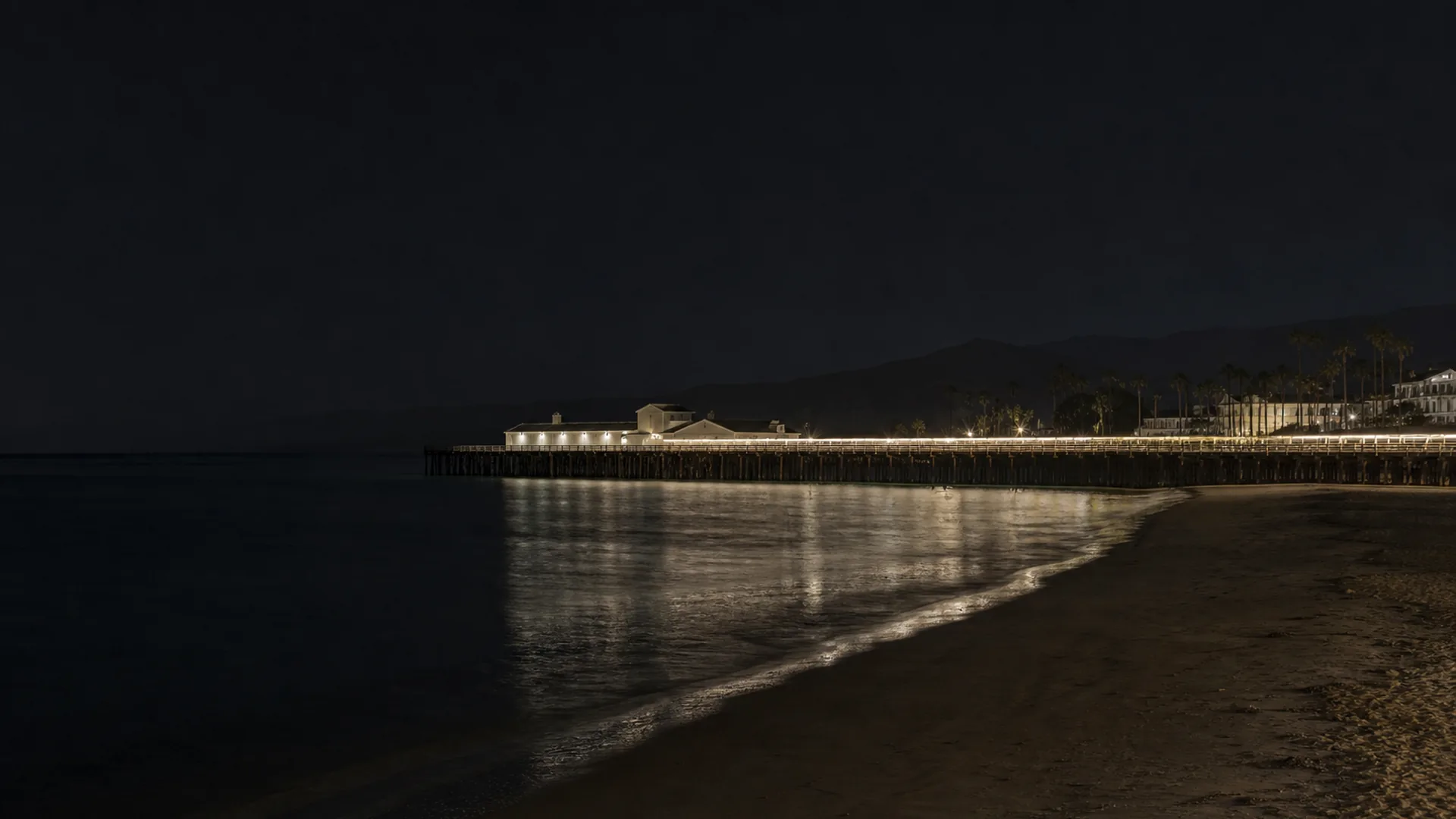 Stearns Wharf extends across dark water from Santa Barbara's beach at blue hour, with the city and mountains beyond.